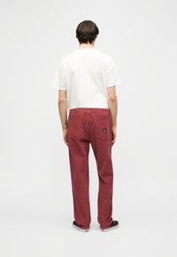 Άνδρας που στέκεται με την πλάτη, φοράει λευκό μπλουζάκι, άνετο κόκκινο παντελόνι με πίσω τσέπες και μαύρα παπούτσια χωρίς κορδόνια.