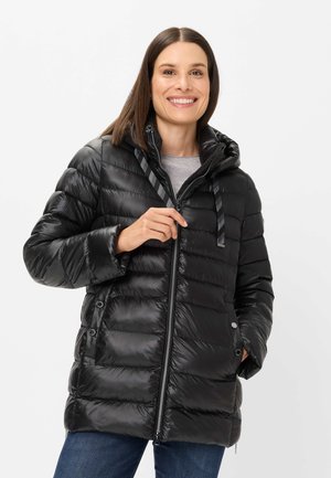 Lächelnde Frau mit glattem braunem Haar, die eine schwarze gesteppte Kapuzenjacke trägt, die teilweise zugezippt ist, mit der linken Hand in der Tasche und der rechten Hand am Reißverschluss.