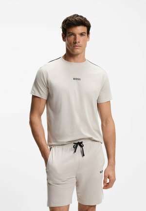 Mężczyzna w jasnobeżowej koszulce BOSS i dopasowanych szortach z czarnym sznurkiem, stoi z jedną ręką w kieszeni na jednolitym tle.