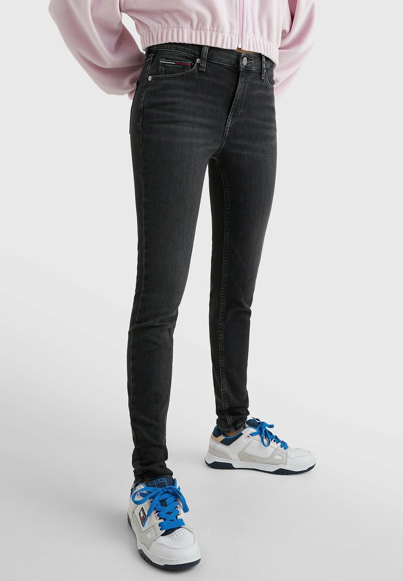 Une personne portant un jean skinny noir, des baskets blanches avec des lacets bleus, et un sweat-shirt court rose se tenant devant un fond blanc uni.