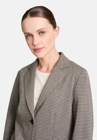 Blazer à carreaux en tissu beige et noir avec une coupe ajustée, revers crantés, fermeture à un bouton et doublure intérieure.