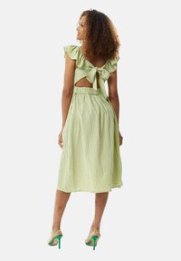 Green and white striped dress with ruffled sleeves, open back, and tied closure. Flared skirt and elastic waistband. Worn with green heels.