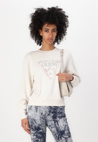 Crème-kleurige sweatshirt met een driehoekig merklogo en strassaccenten, gecombineerd met gestreepte blauwe broek met bloemen- en vogelontwerpen.