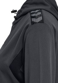 Ciemnoszary, teksturowany rękaw kurtki z czarną naszywką i regulowanym sznurkiem przy ramieniu.