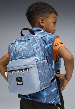 Jeune enfant portant un sac à dos bleu avec un motif d'éclairs et des dents de griffes sur la poche avant, ajustant la sangle en étant de profil.