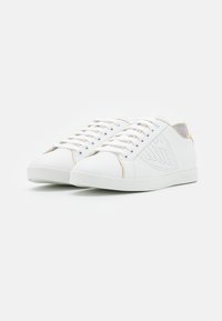 Sneakers bianchi con un esterno in pelle liscia, dettagli dorati, aperture perforate e un design classico con lacci. Presentano una punta arrotondata e una suola piatta.