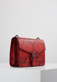 Punainen käärmeenahkainen käsilaukku, jossa on teksturoitu pinta, läppäkiinnitys, metalliosat ja pieni koristeellinen soljen yksityiskohta.