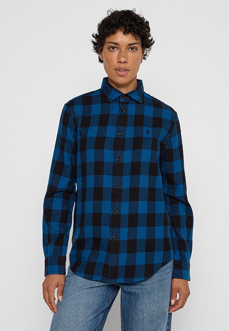 Persona con capelli ricci e corti, che indossa una camicia a maniche lunghe a quadri blu e neri e jeans di un blu chiaro, in piedi su uno sfondo semplice.