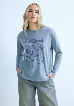 Femme aux longs cheveux blonds portant un chemisier bleu à manches longues avec des illustrations violettes sur le thème de la boulangerie et un pantalon gris, mains dans les poches.