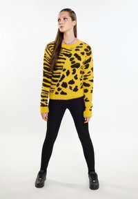 Geel donzig vest met zwart luipaardpatroon, gecombineerd met zwarte skinny jeans en zwarte sneakers. Staande pose met ontspannen handen.