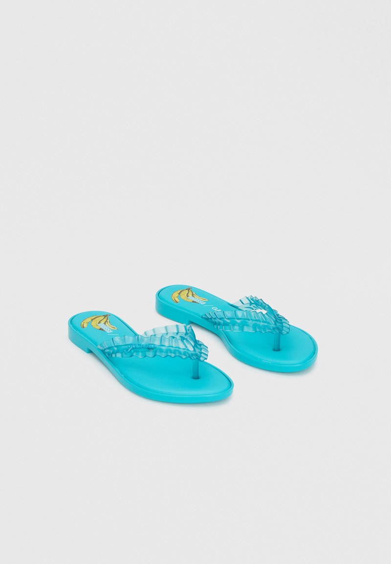 Sandales plates bleues avec des brides à volants et un motif banane sur la semelle, affichées sur fond blanc.