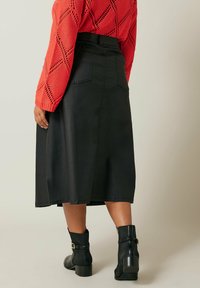 Zwarte middelange rok met een aansluitende taille en twee achterzakken, gecombineerd met een gestructureerde rode trui en zwarte enkellaarzen.