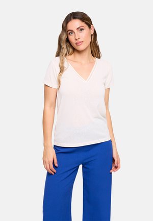 Mujer con cabello largo y ondulado, vestida con una camiseta blanca de manga corta con escote en V y pantalones anchos de color azul brillante, de pie frente a un fondo liso.