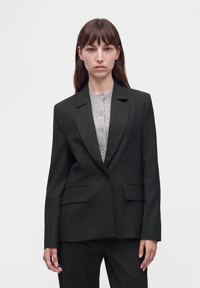 Femme aux cheveux bruns raides porte une veste de costume noire et une chemise grise boutonnée, debout devant un fond blanc uni.