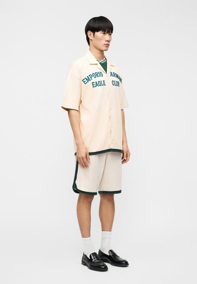 Νεαρός άνδρας με μπεζ πουκάμισο με κουμπιά που έχει το κείμενο «Emporio Armani Eagle Club», ταιριαστά σορτς με σκούρες λεπτομέρειες, λευκές κάλτσες και μαύρα μοκασίνια, στέκεται πλευρικά.
