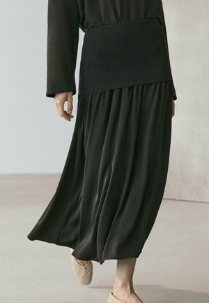 Person trägt einen fließenden schwarzen Rock, ein passendes Oberteil und beige Ballerinas, geht drinnen auf einem hellfarbenen Boden.