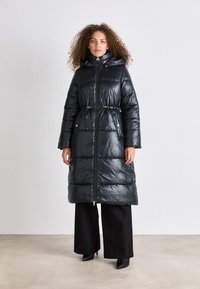 Casaco puffer longo em material preto brilhante, com fecho em zipper à frente, capuz, cintura com cordão e detalhes em ouro. Usado sobre calças largas pretas.