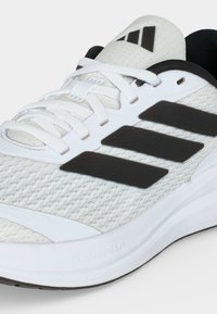 Chaussure de sport blanche avec des bandes noires, tige en mesh, lacets blancs et semelle coussinée marquée "Cloudfoam" sur un fond uni.