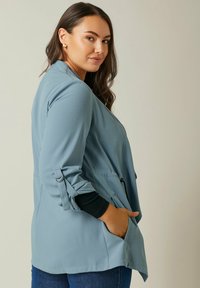 Vaaleansininen bleiseri, jossa on tekstuuroitu kangas, auki oleva etuosa, sivutaskut ja käännetyt hihat, joissa on metalliset yksityiskohdat.
