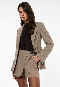 Brun hunternet blazer over en mørk top, parret med matchende skræddersyede shorts med bælte. Struktureret stof med et subtilt ternet mønster.