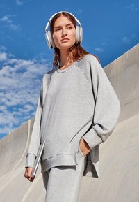 Grijze oversized sweatshirt met raglanmouwen, zijsplitten en een zachte textuur, gedragen met bijpassende broeken. Model houdt een laptop vast.