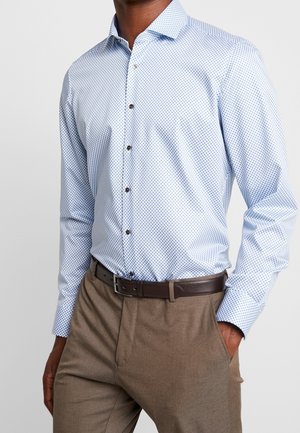 Man wearing light blue patterned long-sleeve shirt tucked into brown trousers with a dark brown leather belt, hand in pocket.