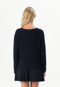 Femeie cu păr blond, purtând un pulover lung cu mânecă lungă de culoare bleu marin și o fustă plisată neagră, stând cu spatele față de un fundal alb.