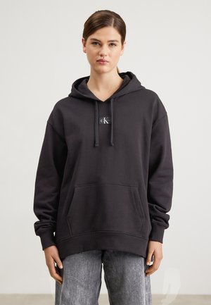Sweatshirt à capuche noire en tissu doux, avec une poche avant, des poignets côtelés et un petit patch logo au centre.