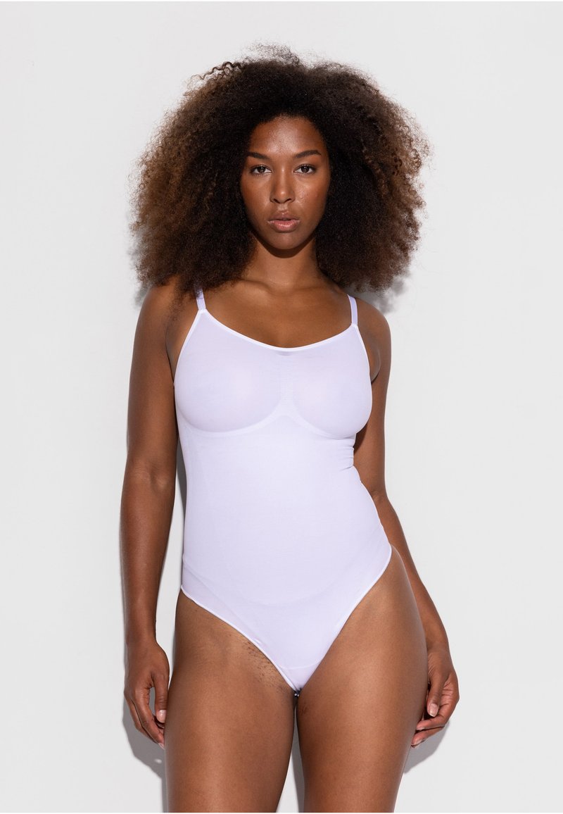 Femme aux cheveux bouclés naturels portant un body blanc ajusté à fines bretelles, debout devant un fond clair uni.