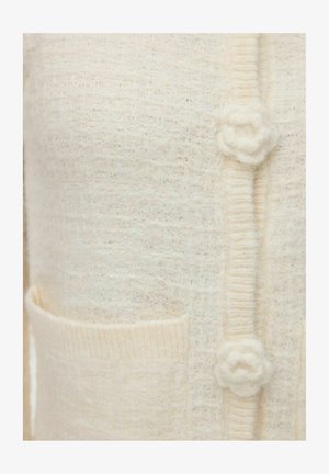 Pleteni kremni kardigan z teksturirano tkanino, ki ima majhen gumb v obliki cveta in sprednji žep. Nežna, topla zasnova.