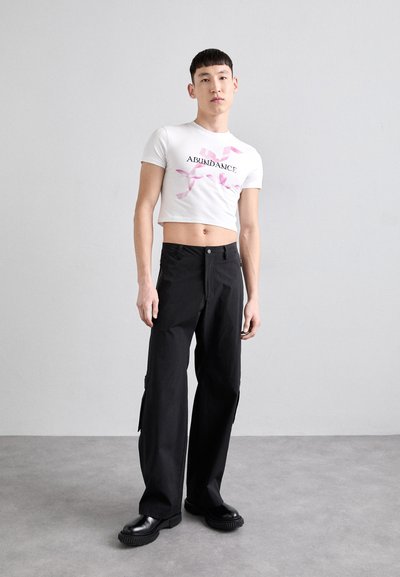 T-shirt blanc court avec le texte "ABONDANCE" et un graphique de ruban rose, associé à un pantalon cargo noir taille haute et des chaussures compensées noires.