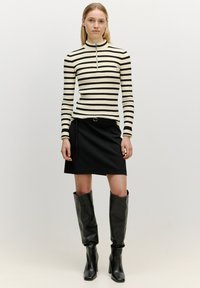 Schwarz-weiß gestreifter ribbeliger Pullover mit hohem Kragen und Reißverschlussdetail, kombiniert mit einem schwarzen Rock und knielangen schwarzen Lederstiefeln.
