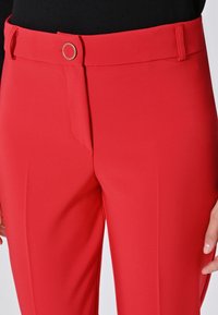 Pantaloni rossi a vita alta con chiusura a bottone circolare argentato, indossati con una maglietta nera, che mostrano parte della mano destra su uno sfondo neutro.