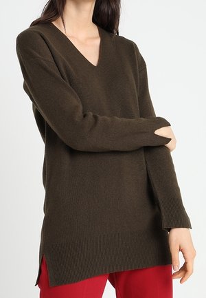 Donna che indossa un maglione lungo a V di colore marrone scuro con orli e polsini a coste, abbinato a pantaloni rossi, in posa contro uno sfondo bianco uniforme.