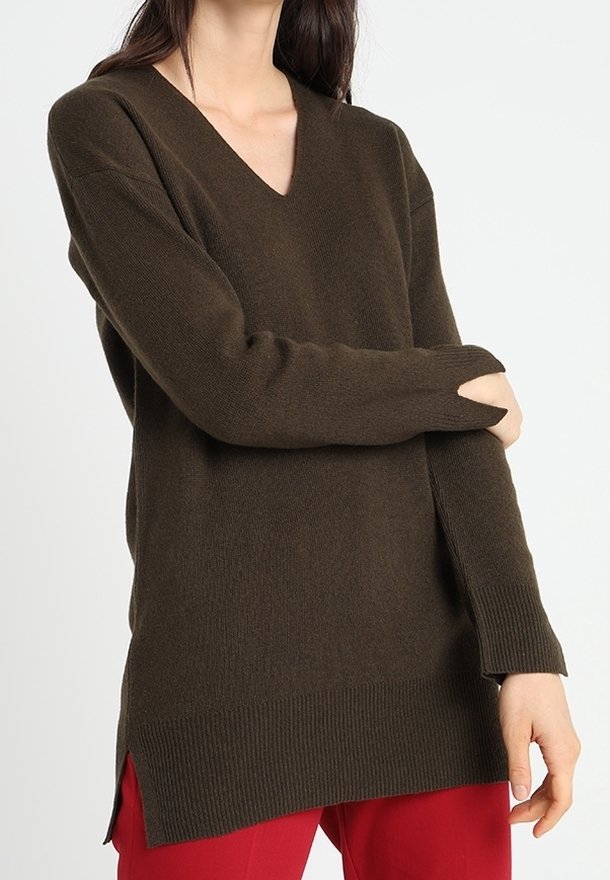 Donna che indossa un maglione lungo a V di colore marrone scuro con orli e polsini a coste, abbinato a pantaloni rossi, in posa contro uno sfondo bianco uniforme.