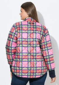 Kvinde iført en farverig quiltet jakke med et plaidmønster i pink, grøn, orange og blå, set fra ryggen mod en hvid baggrund.