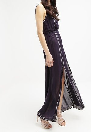 Femme portant une robe longue foncée sans manches, avec bordure blanche et fente devant, associée à des sandales à talons hauts de couleur claire, debout de côté.