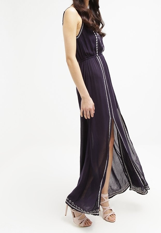 Femme portant une robe longue foncée sans manches, avec bordure blanche et fente devant, associée à des sandales à talons hauts de couleur claire, debout de côté.