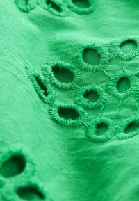 Tessuto verde con cerchi ritagliati ricamati. La superficie è liscia con cuciture in rilievo attorno alle aperture, creando un design a motivo.