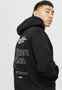 Haut à capuche noir avec un grand motif au dos. Le design du texte comprend "Runners Assoc." et un texte supplémentaire en blanc. Tissu doux, coupe décontractée.