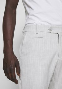 Dark-skinned person wearing light gray textured trousers with a button and zipper, paired with a white shirt, hand relaxed by their side.
