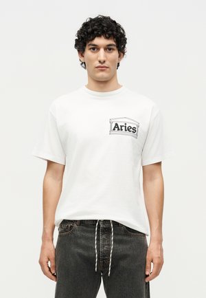 Junger Mann mit lockigem Haar, der ein weißes T-Shirt mit dem Schriftzug "Aries" und schwarze Jeans mit weißem Kordelzug trägt, steht vor einem schlichten Hintergrund.