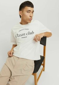 Balta medvilninė marškinė su juodu tekstu "Amalfi Coast Ocean club PREMIUM." Atsitiktinis siluetas, apvalus iškirpimas, trumpomis rankovėmis. Šviesiai rudos kelnės.