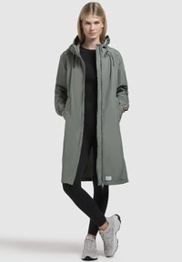 Jachetă lungă, de culoare verde deschis, cu glugă, închidere cu fermoar și buzunare laterale, purtată peste un top negru și colanți negri. Material texturat, neted.