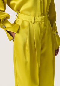 Blusa gialla in satin con maniche lunghe e polsini con bottoni, con accessori decorativi. Pantaloni a vita alta e plissettati in tessuto coordinato.