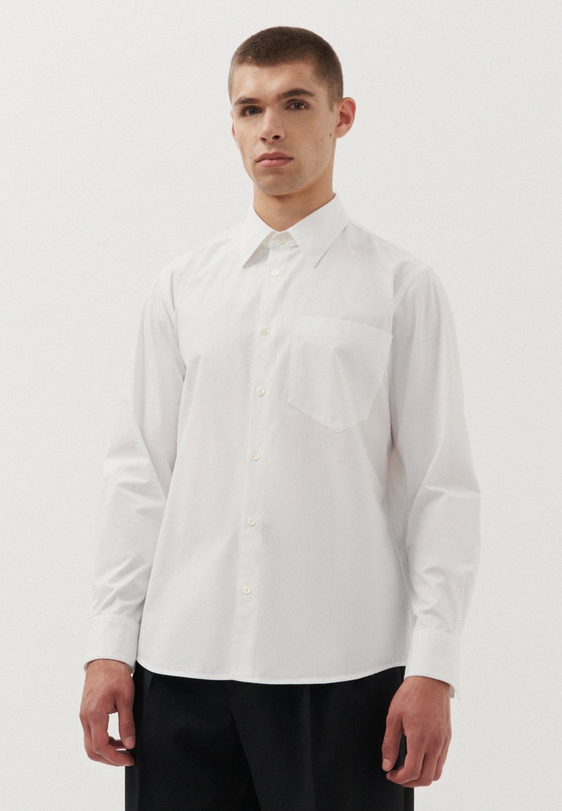 Jeune homme aux cheveux courts portant une chemise blanche à manches longues boutonnée et un pantalon noir devant un fond clair uni.