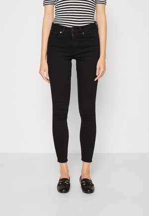 Nainen, joka pukeutunut mustiin skinny-farkkuihin, mustiin loafer-kenkiin ja mustavalkoraidalliseen lyhythihaiseen toppiin, seisoo yksivärisen valkoisen taustan edessä.