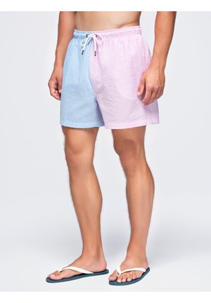 Homme portant un short texturé bicolore, côté gauche bleu clair, côté droit rose clair, assorti de tongs blanches sur un fond uni.