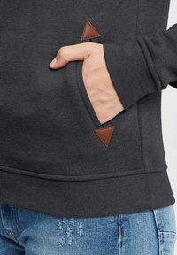 Tmavě šedá mikina s texturovanou tkaninou, která má přední kapsu s hnědým koženým trojúhelníkovým detailem. Nosí se s popraskanými modrými džíny.