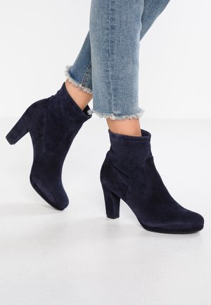 Kvinde iført marineblå ruskindsstøvler med hæl, kombineret med cropped lyseblå jeans med frynset kant, stående på en ensfarvet hvid gulv.